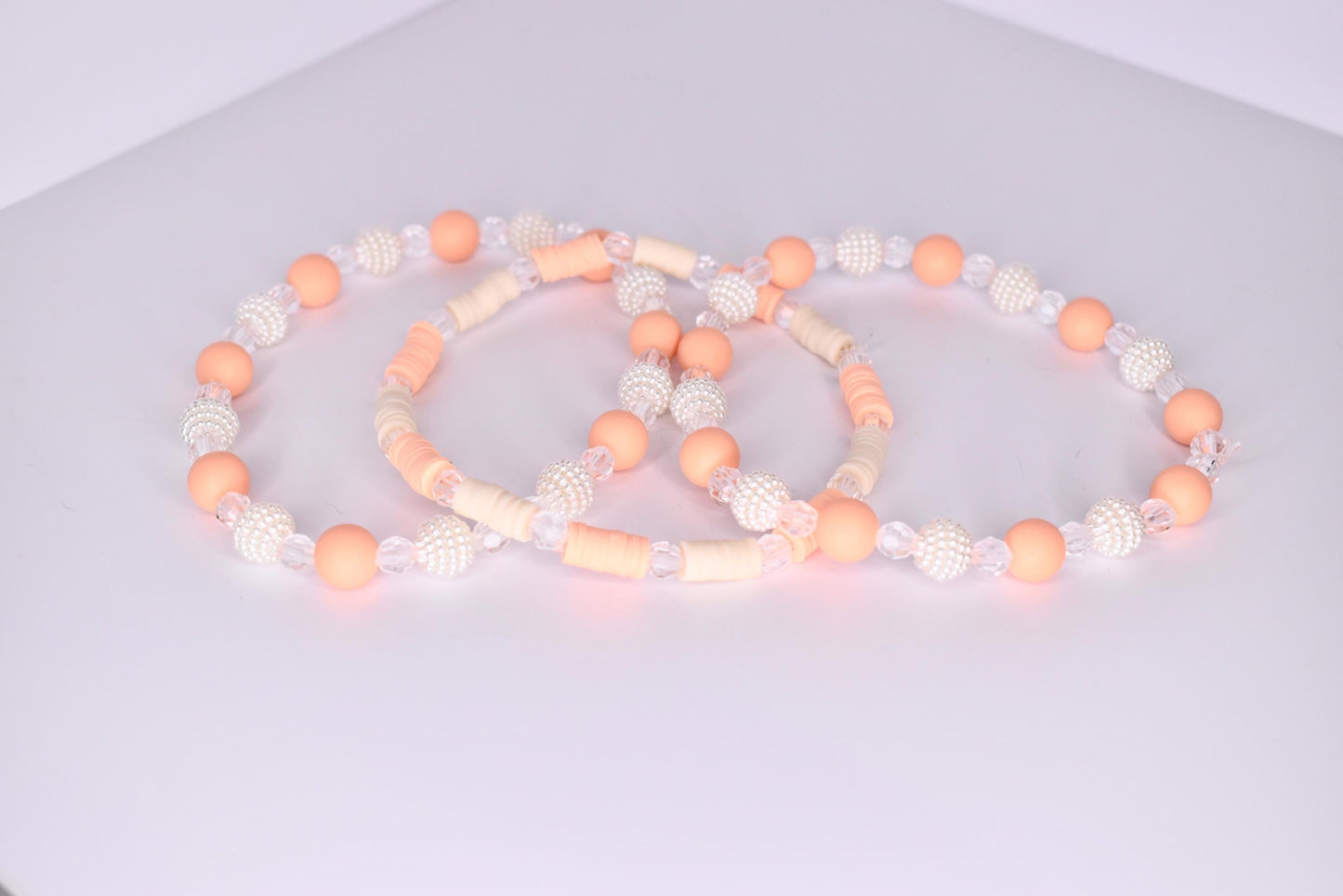 A trio of tumbler bracelets arranged in an overlapping display on a white surface, featuring a mix of peach matte beads, clear faceted beads, textured white pearl beads, and flat cream and peach disc beads in complementary patterns.
