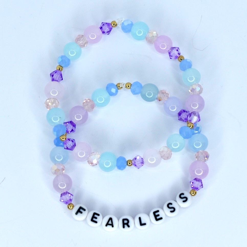 Two beaded bracelets in shades of blue, purple, and pink. The bracelets feature round and faceted beads, with gold accents. The bottom bracelet spells 'FEARLESS' in white letter beads. The bracelets are likely fashion accessories.