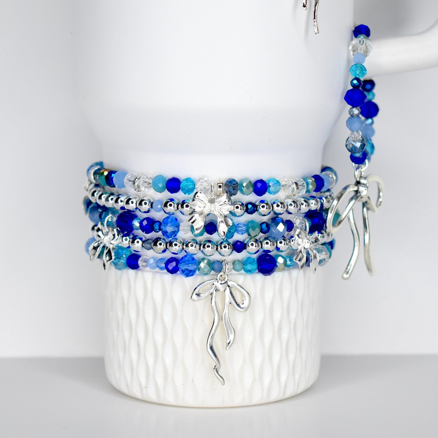 A collection of blue beaded bracelets with silver accents and charms. The bracelets feature various shades of blue beads, including light blue, dark blue, and clear beads. Silver bow-shaped charms dangle from the bracelets. The bracelets are displayed on a white Stanley.