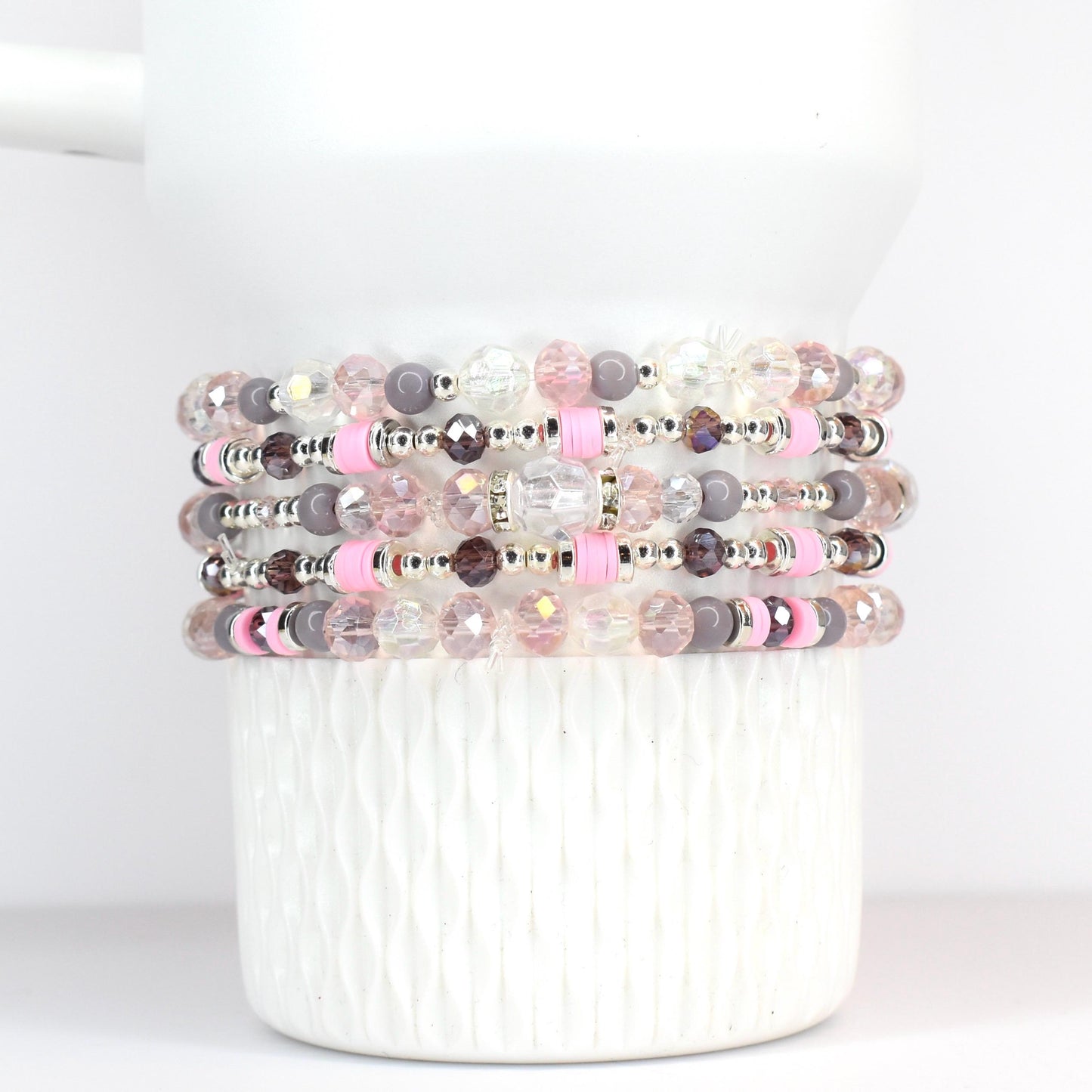 Stack of beaded bracelets in shades of pink, gray, and clear. The bracelets feature a variety of beads, including faceted crystal, round gray beads, and pink spacers. The bracelets are stacked on a white, textured surface.