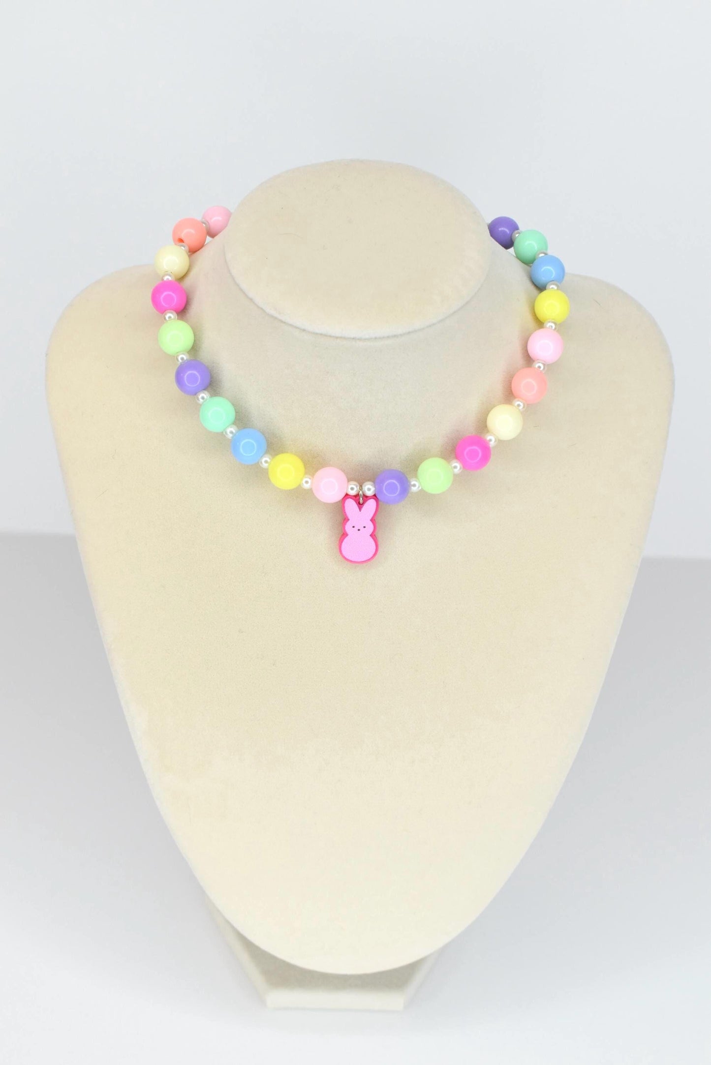 A colorful beaded necklace with pastel-colored beads in pink, yellow, green, blue, and purple. A pink bunny charm hangs from the center. The necklace is displayed on a mannequin.