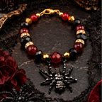 A gothic-style beaded tumbler handle bracelet with red and black faceted beads, gold-toned spacer rings, and a black spider charm embellished with rhinestones, displayed on a dark textured surface with Halloween-themed props.