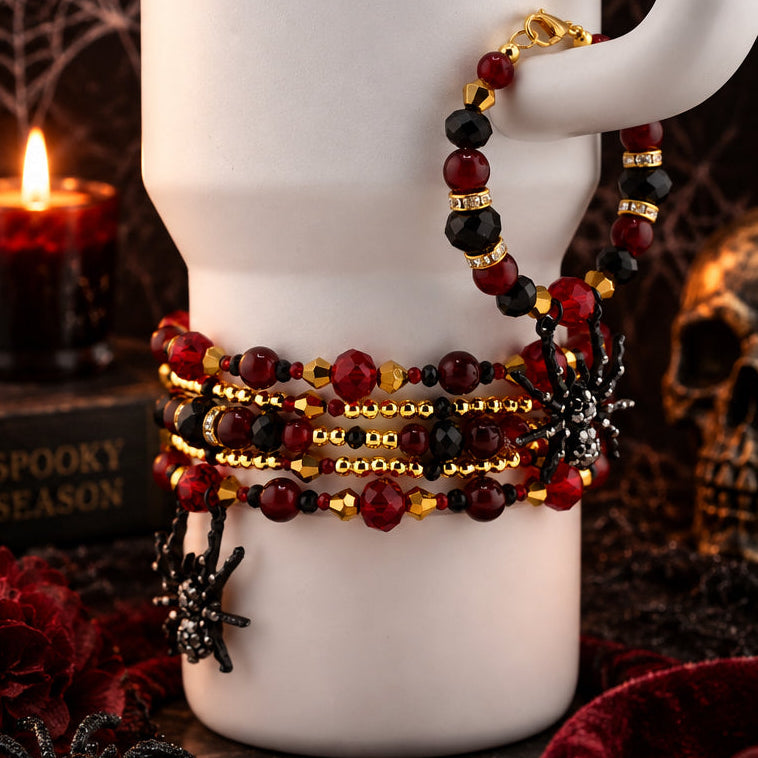 A set of five beaded stretch bracelets with red, black, and gold beads, spider charms, and a black spiderweb clasp, displayed on a white ceramic mug with a gothic Halloween background.