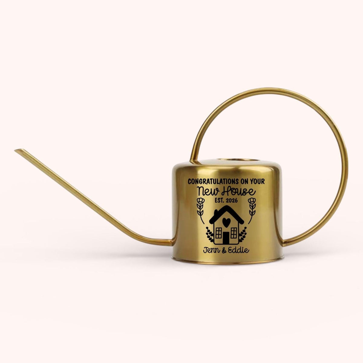 A gold-colored watering can with a long spout and a circular handle. The can features the text "Congratulations on your New House" with a house graphic and the names "Jenn & Eddie".