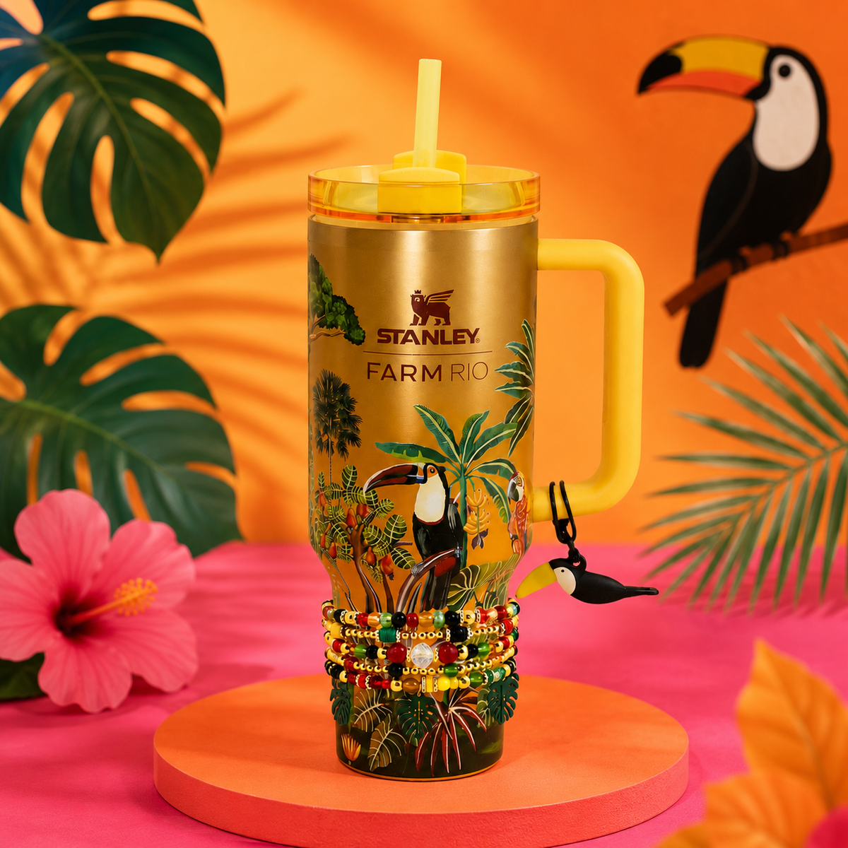 Stanley Farm Rio insulated tumbler with a yellow handle and lid, decorated with tropical jungle artwork and toucan motifs, displayed on a pink surface with tropical props and a bright orange background.