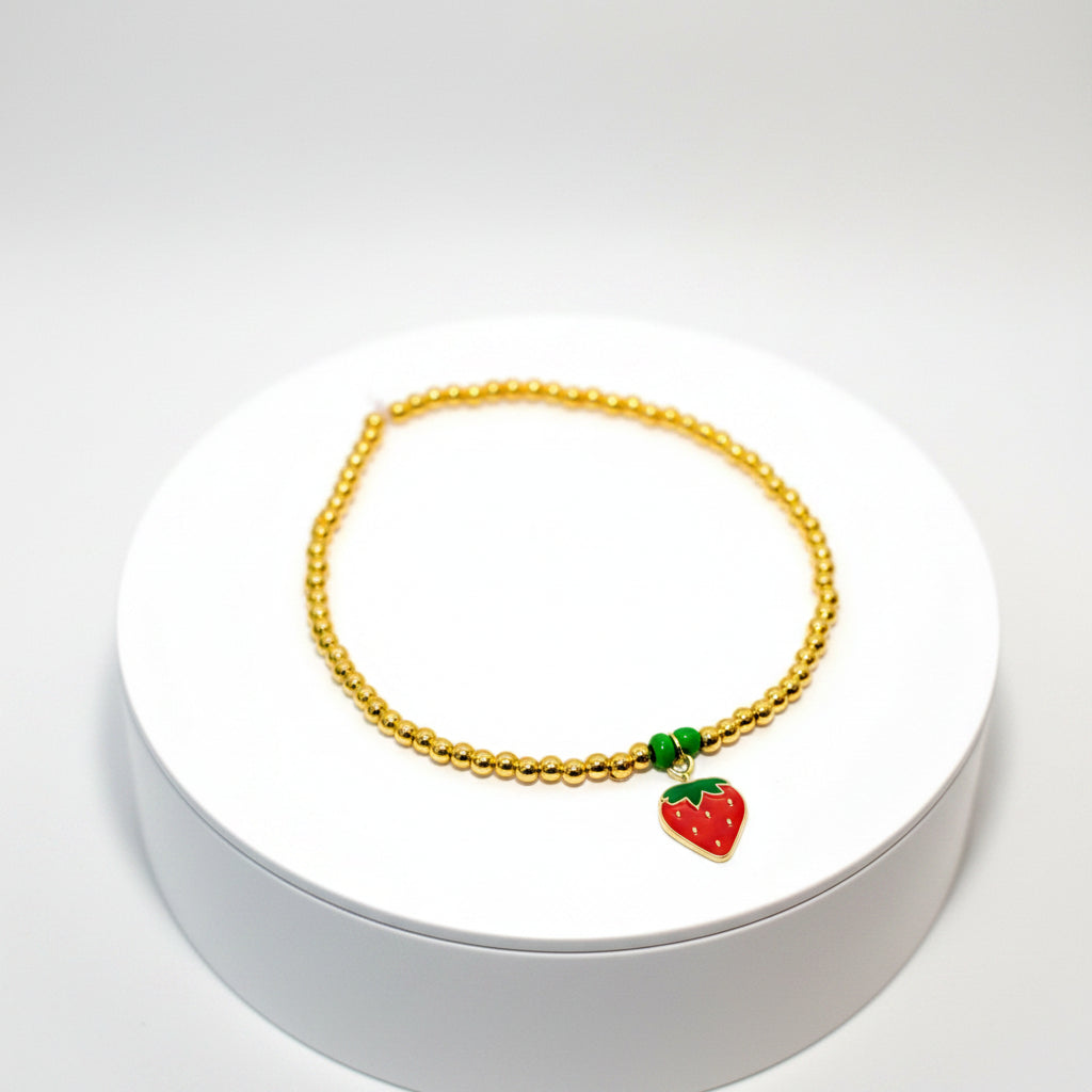 A gold beaded bracelet with a red and green strawberry charm. The bracelet has a design name of Strawberry Kiss.