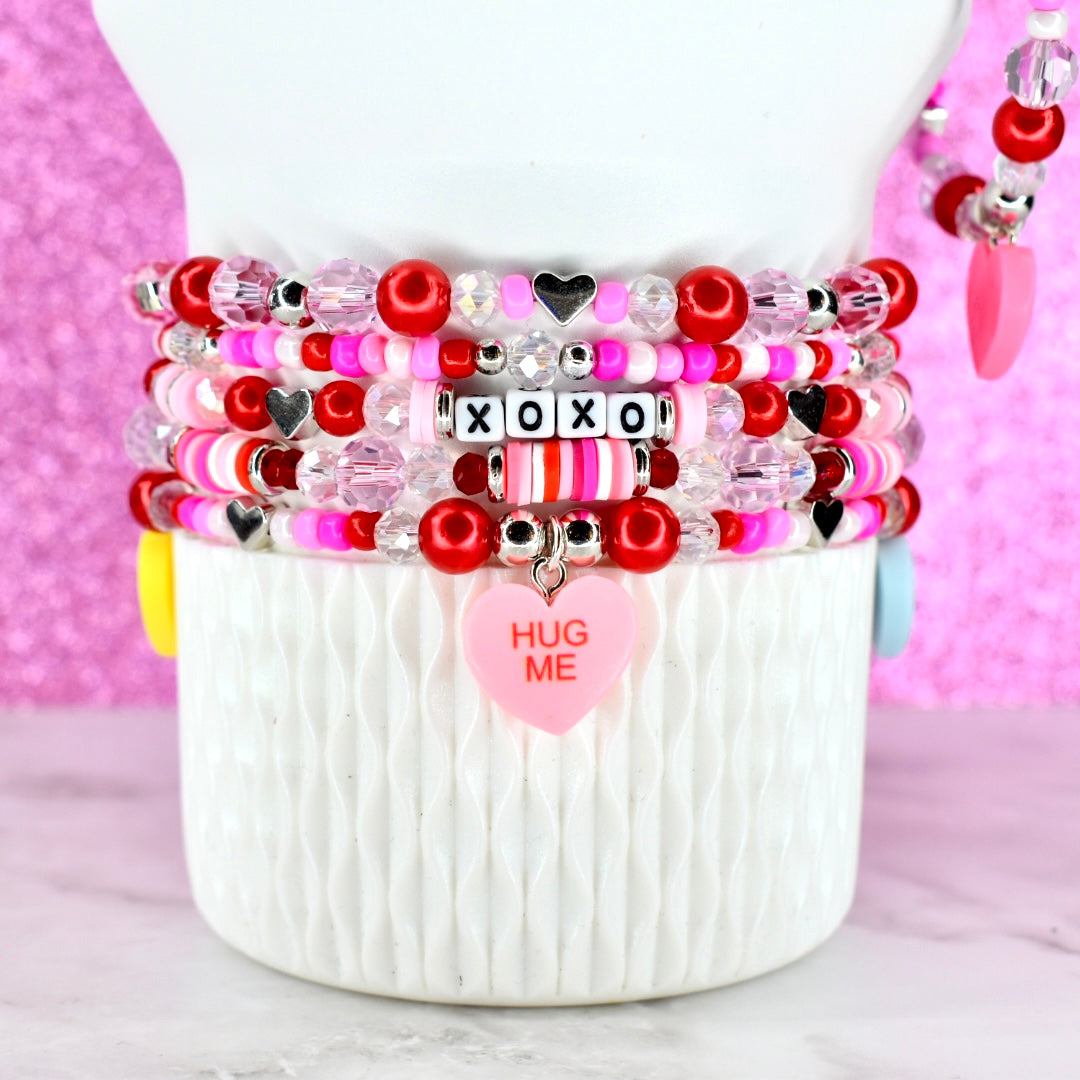 Stack of colorful beaded bracelets with heart and 'Hug Me' charms on a white Stanley tumbler against a pink background.