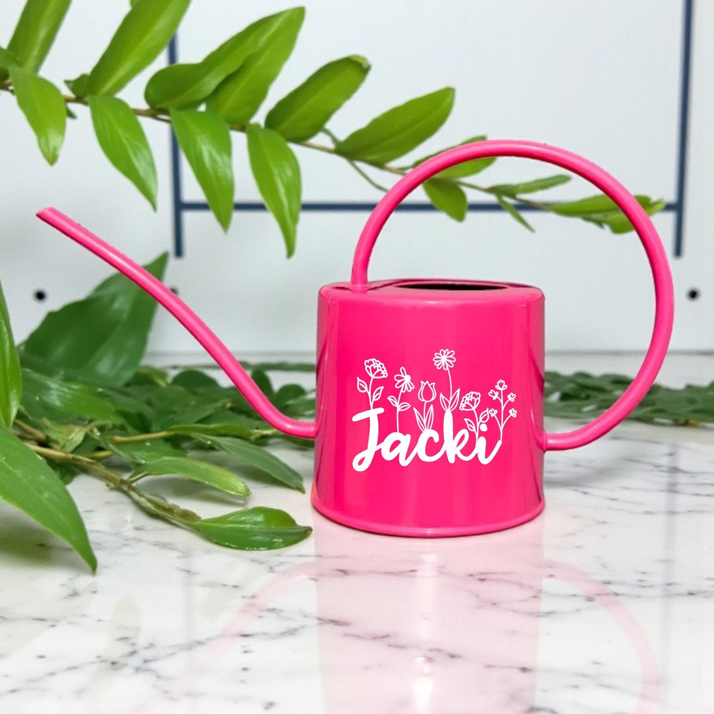 Pink watering can with a name on a marble surface with green leaves