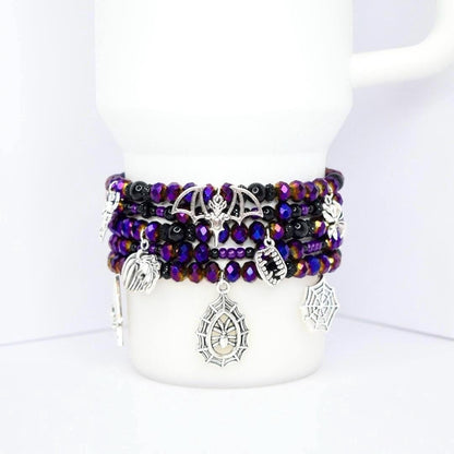 Calathea Collective gothic Halloween charm bracelet stack with purple, black, and gold beads and silver bat, spider web, and fangs charms.