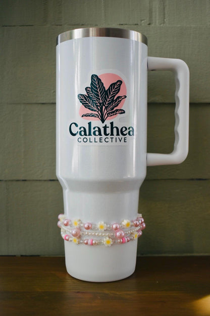 White travel mug with a pink and green logo that says "Calathea Collective". The mug has a white handle and is decorated with a pink and white beaded band.