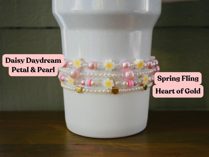 A white tumbler with four layers of pink and white beaded bracelets. The bracelets have small pink flowers and gold heart charms. The text "Daisy Daydream Petal & Pearl" is on the left and "Spring Fling Heart of Gold" is on the right.