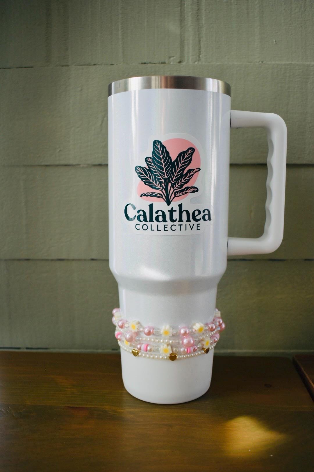 White stainless steel travel mug with a black and pink logo that says "Calathea Collective" and a green plant design. The mug has a handle and is decorated with a pink and white beaded bracelet.
