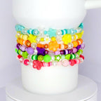 A stack of colorful beaded bracelets. The bracelets feature various shapes, including stars and flowers, and a range of colors like pink, green, yellow, and purple. The bracelets are stacked on a white Stanley tumbler.