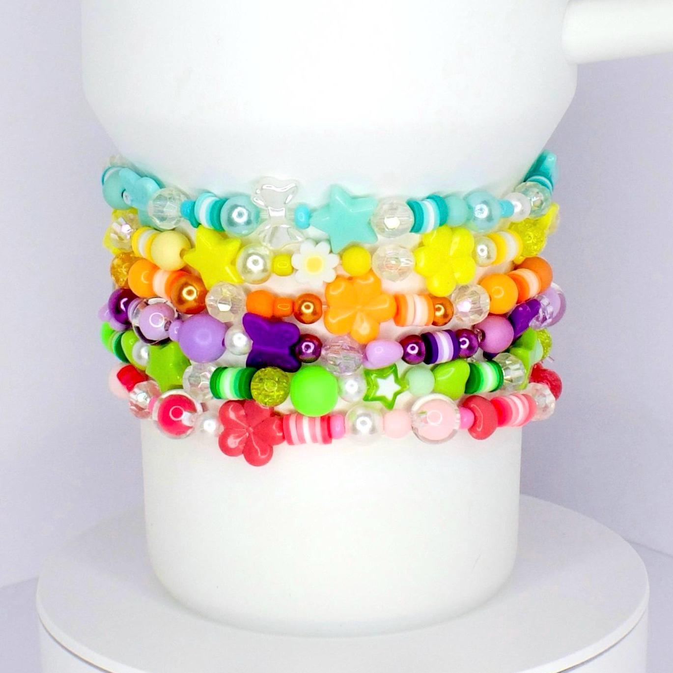 A stack of colorful beaded bracelets. The bracelets feature various shapes, including stars and flowers, and a range of colors like pink, green, yellow, and purple. The bracelets are stacked on a white Stanley tumbler.