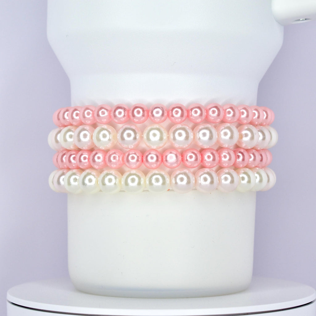 A white Stanley tumbler wrapped with a classic and elegant bracelet stack made of alternating rows of glossy pink and white pearl beads. The bracelets are styled as Stanley cup accessories with a soft and feminine touch.