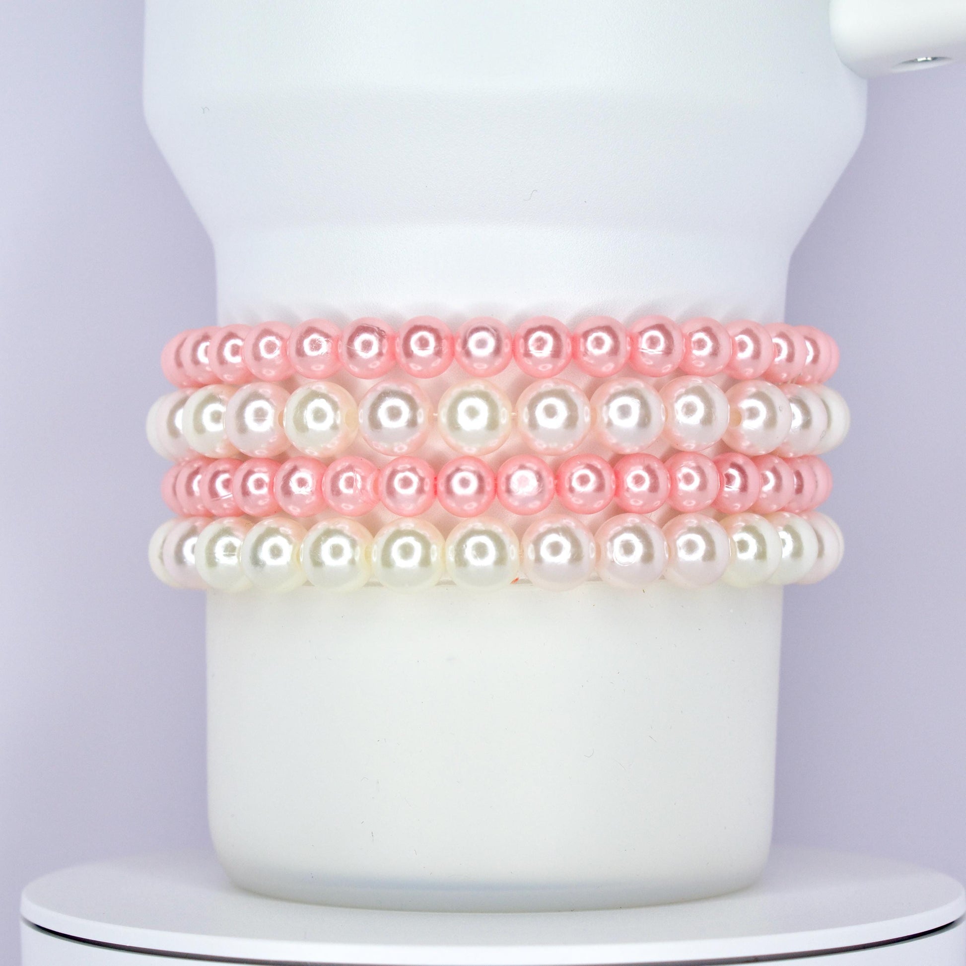 A white Stanley tumbler wrapped with a classic and elegant bracelet stack made of alternating rows of glossy pink and white pearl beads. The bracelets are styled as Stanley cup accessories with a soft and feminine touch.