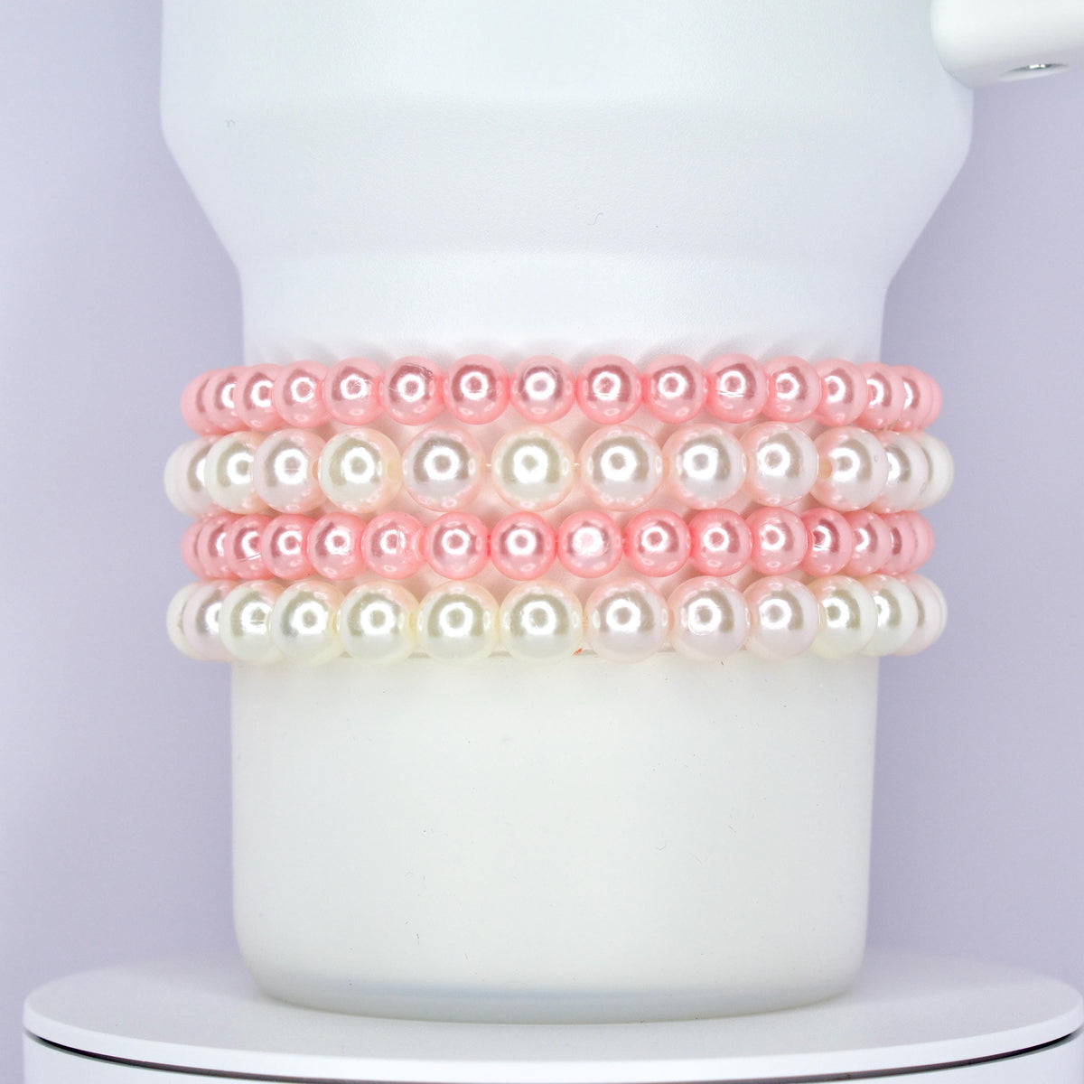 A white Stanley tumbler wrapped with a classic and elegant bracelet stack made of alternating rows of glossy pink and white pearl beads. The bracelets are styled as Stanley cup accessories with a soft and feminine touch.