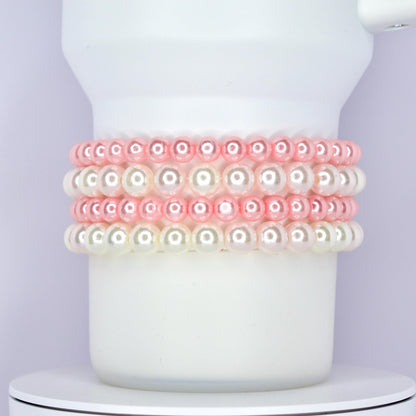 A white Stanley tumbler wrapped with a classic and elegant bracelet stack made of alternating rows of glossy pink and white pearl beads. The bracelets are styled as Stanley cup accessories with a soft and feminine touch.