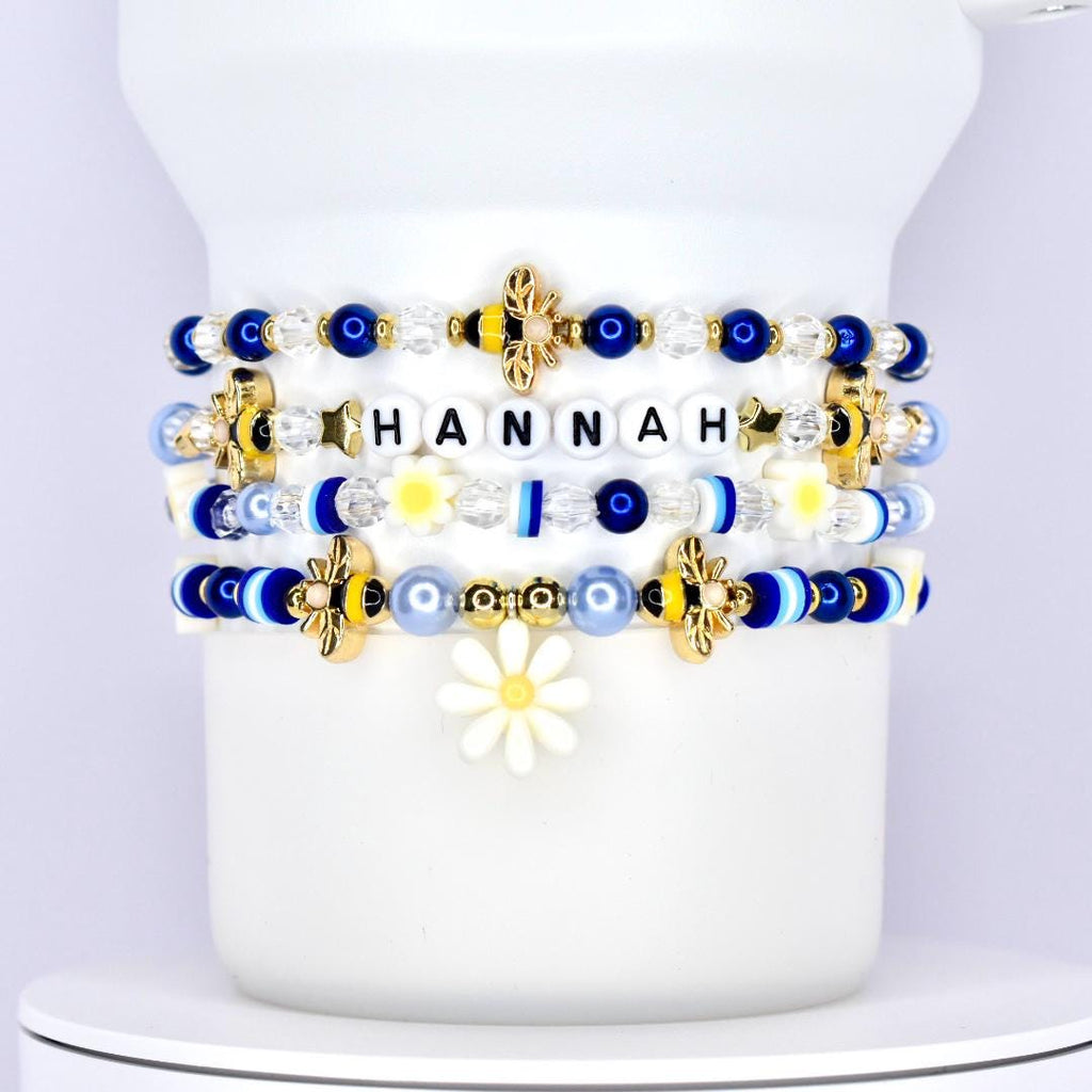 A stack of five custom tumbler bracelets wrapped around a white tumbler, featuring a personalized name bracelet with the name "HANNAH" in black letter beads, and decorated with a mix of blue, white, gold, and yellow beads, including gold bees, daisies, smiley faces, stars, and striped accent beads in various textures and finishes.