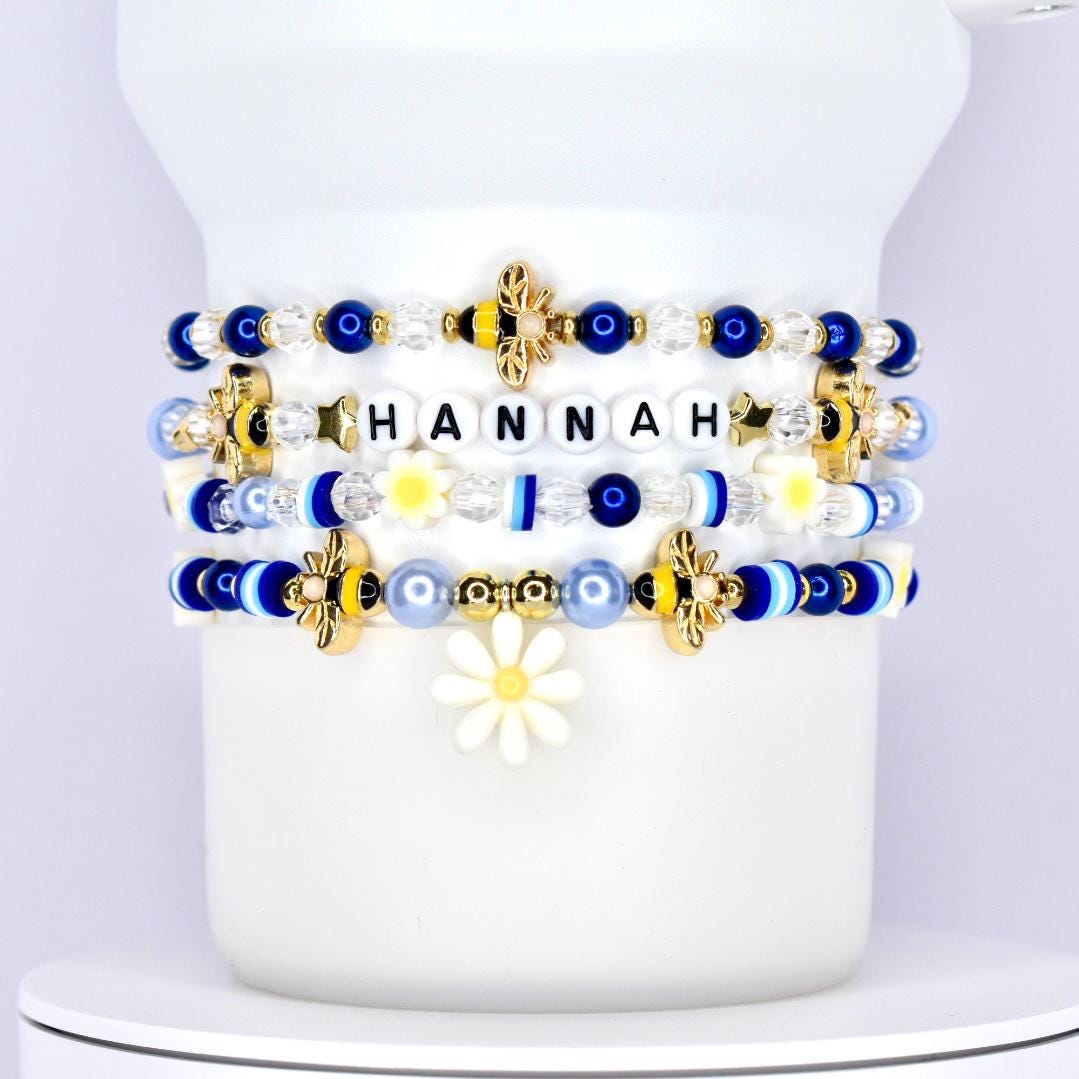 A stack of five custom tumbler bracelets wrapped around a white tumbler, featuring a personalized name bracelet with the name "HANNAH" in black letter beads, and decorated with a mix of blue, white, gold, and yellow beads, including gold bees, daisies, smiley faces, stars, and striped accent beads in various textures and finishes.