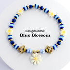 A blue and white beaded bracelet with a gold bee charm and a white daisy charm. The bracelet has a blue and white striped pattern and is decorated with small white flowers. The text 'Design Name: Blue Blossom' is printed below the bracelet.