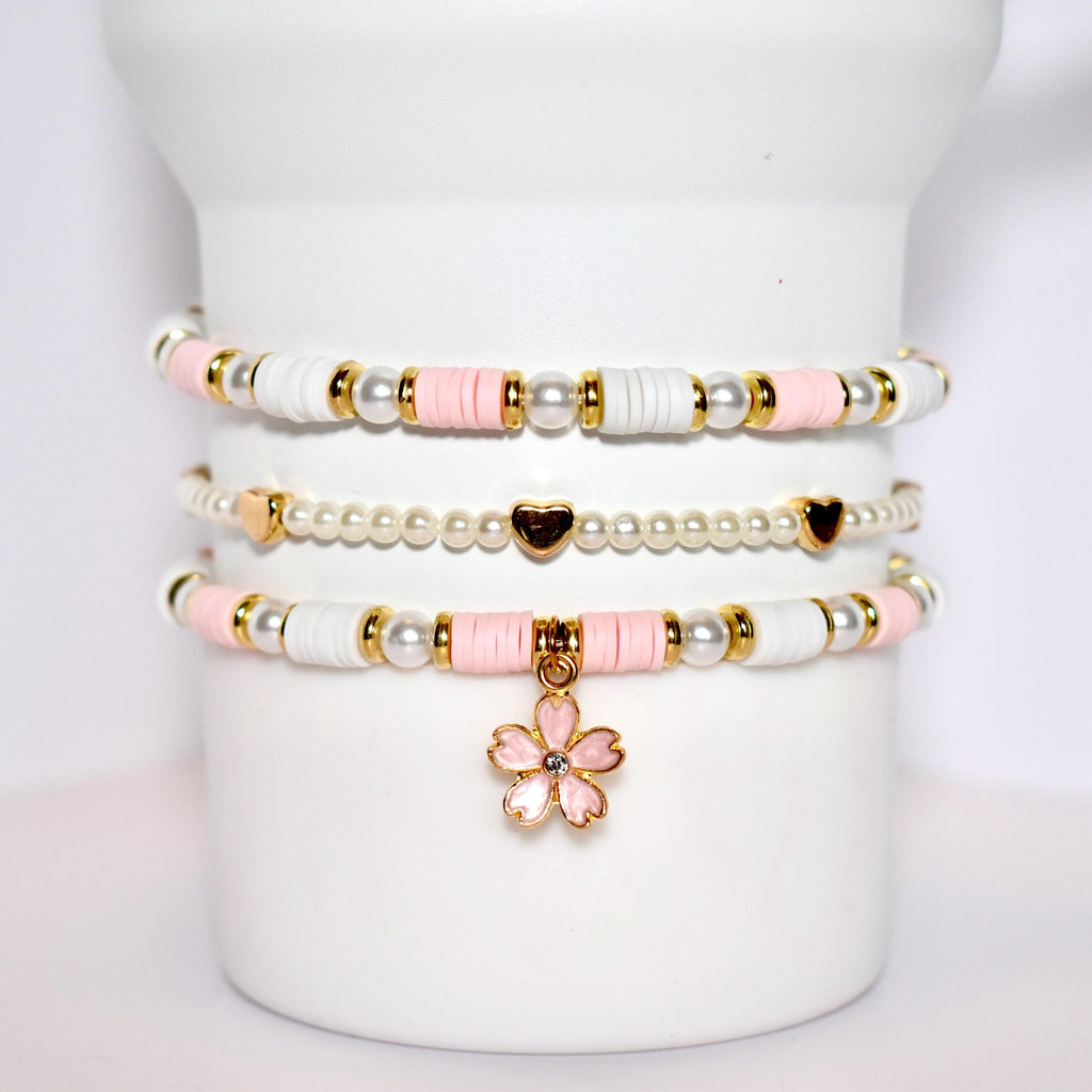 A set of three bracelets with pink and white beads and gold accents. The bracelets are stacked on top of each other and have a pink flower charm hanging from the bottom.