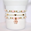 A set of three bracelets with pink and white beads and gold accents. The bracelets are stacked on top of each other and have a pink flower charm hanging from the bottom.