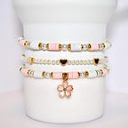 A set of three bracelets with pink and white beads and gold accents. The bracelets are stacked on top of each other and have a pink flower charm hanging from the bottom.