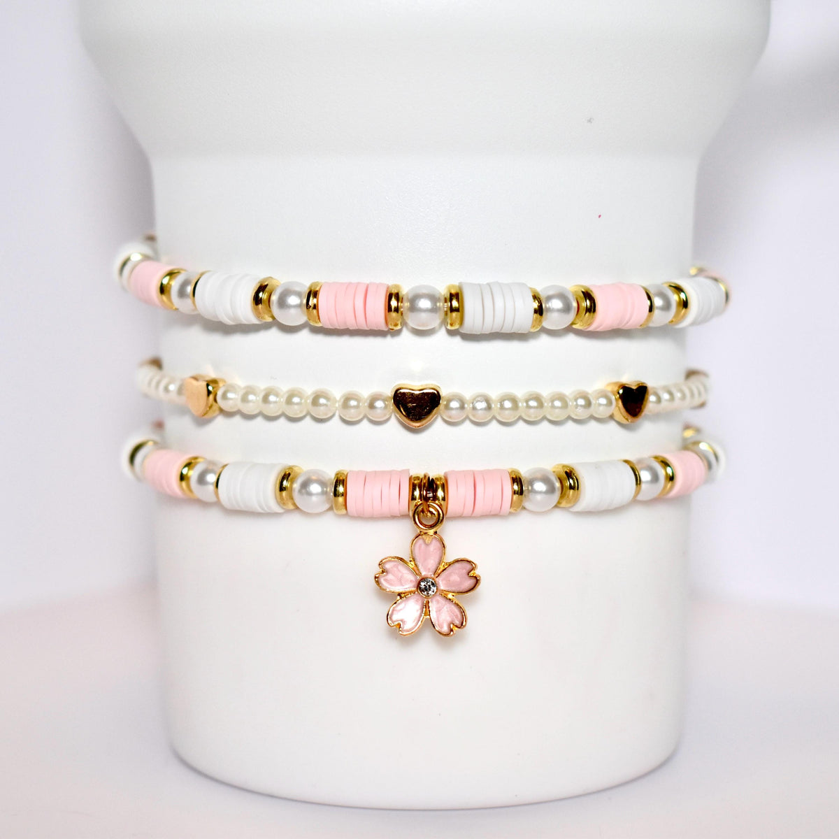 A set of three bracelets with pink and white beads and gold accents. The bracelets are stacked on top of each other and have a pink flower charm hanging from the bottom.