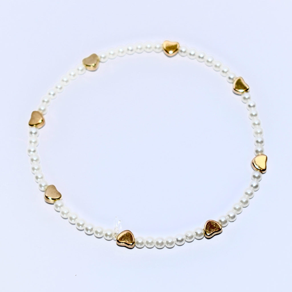 A white pearl bracelet with gold heart charms. The bracelet is made of small, white pearls and has seven gold heart charms evenly spaced around the bracelet.