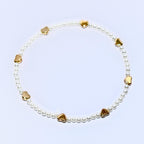 A white pearl bracelet with gold heart charms. The bracelet is made of small, white pearls and has seven gold heart charms evenly spaced around the bracelet.