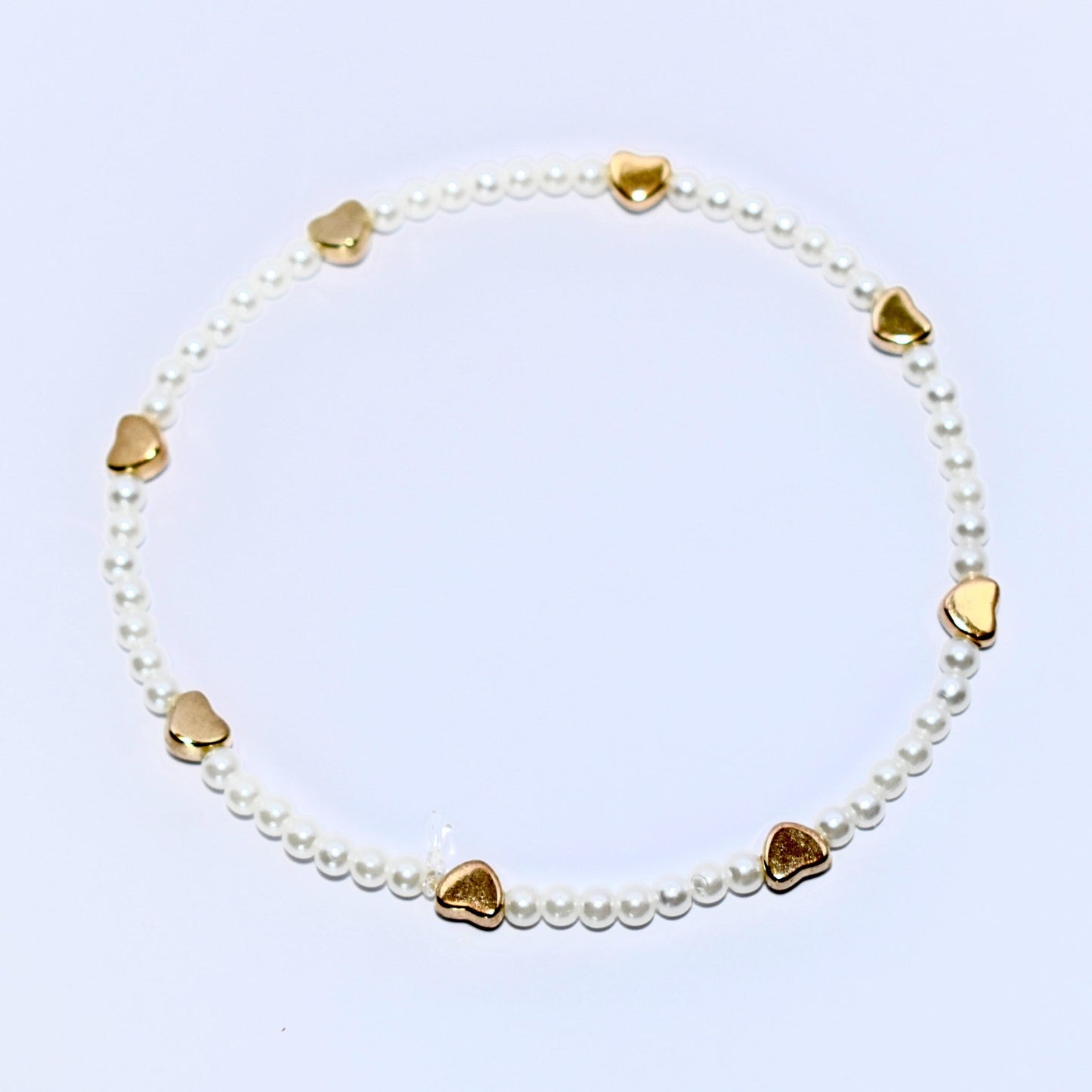 A white pearl bracelet with gold heart charms. The bracelet is made of small, white pearls and has seven gold heart charms evenly spaced around the bracelet.