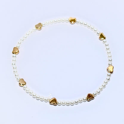 A white pearl bracelet with gold heart charms. The bracelet is made of small, white pearls and has seven gold heart charms evenly spaced around the bracelet.