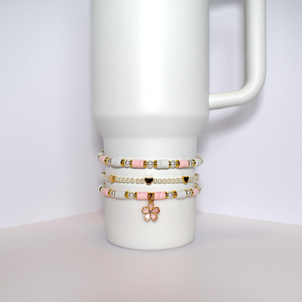 Three beaded bracelets stacked on top of each other on a white mug. The bracelets are made of white and pink beads with gold accents. The top bracelet has a gold flower charm.