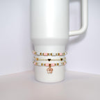 Three beaded bracelets stacked on top of each other on a white mug. The bracelets are made of white and pink beads with gold accents. The top bracelet has a gold flower charm.