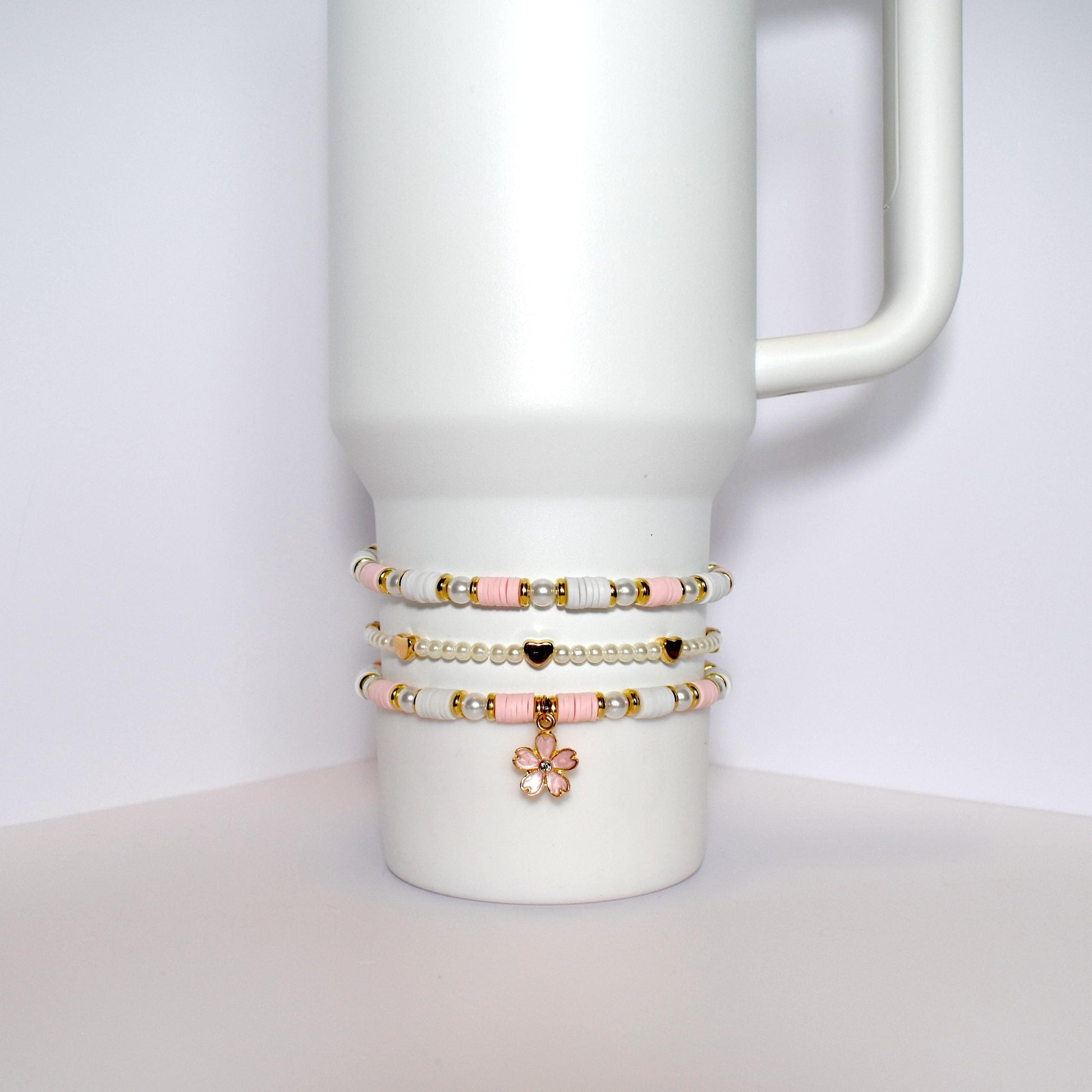 Three beaded bracelets stacked on top of each other on a white mug. The bracelets are made of white and pink beads with gold accents. The top bracelet has a gold flower charm.