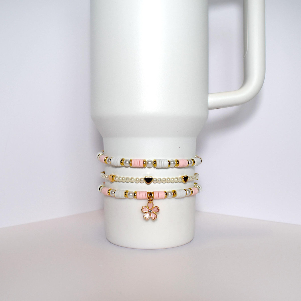 Three beaded bracelets stacked on top of each other on a white mug. The bracelets are made of white and pink beads with gold accents. The top bracelet has a gold flower charm.