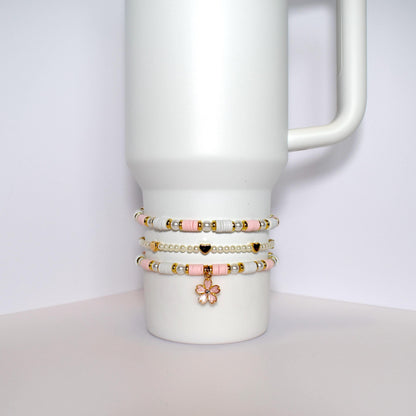 Three beaded bracelets stacked on top of each other on a white mug. The bracelets are made of white and pink beads with gold accents. The top bracelet has a gold flower charm.