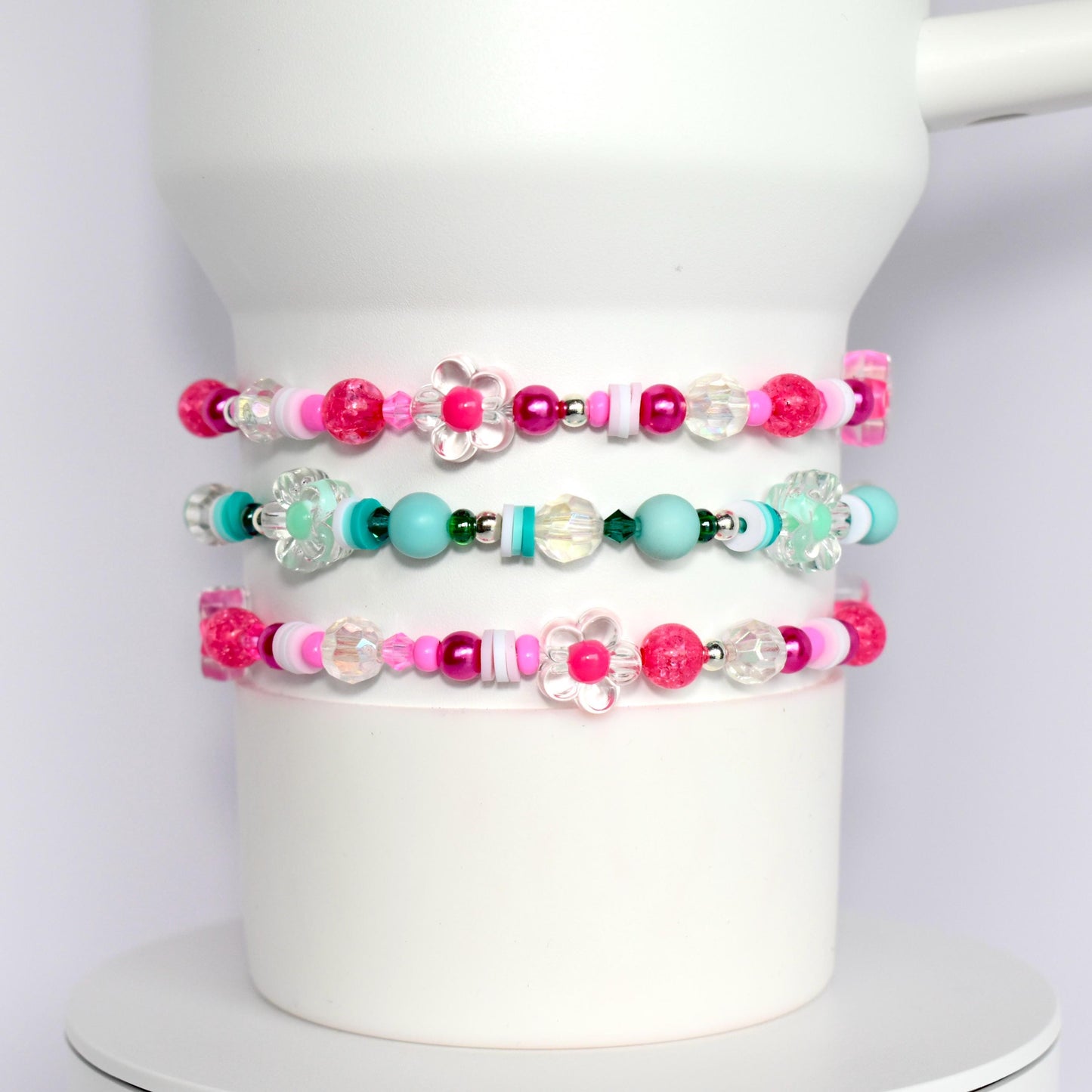 Three pink, white, and turquoise beaded bracelets with flower charms. The bracelets are stacked on top of each other.