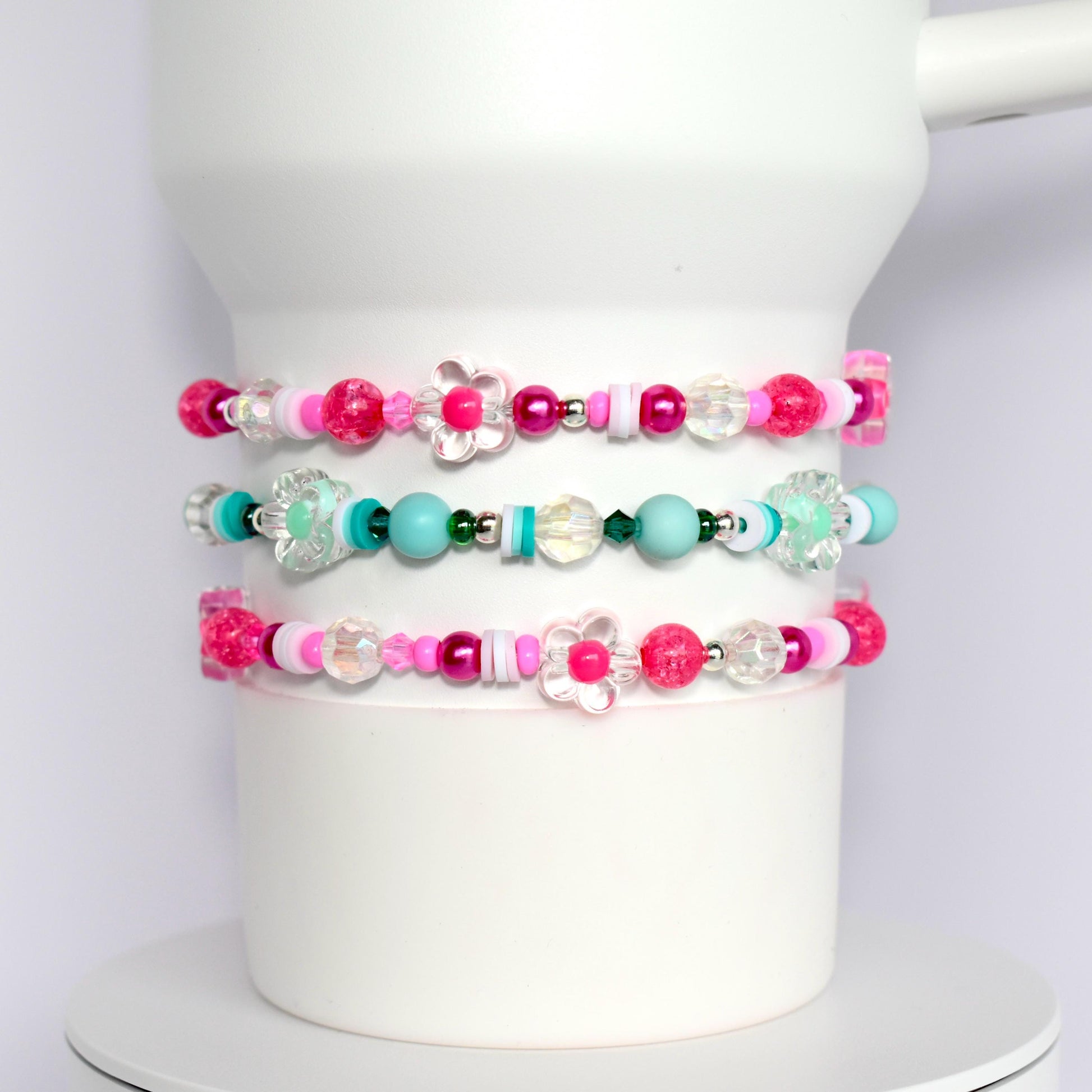 Three pink, white, and turquoise beaded bracelets with flower charms. The bracelets are stacked on top of each other.