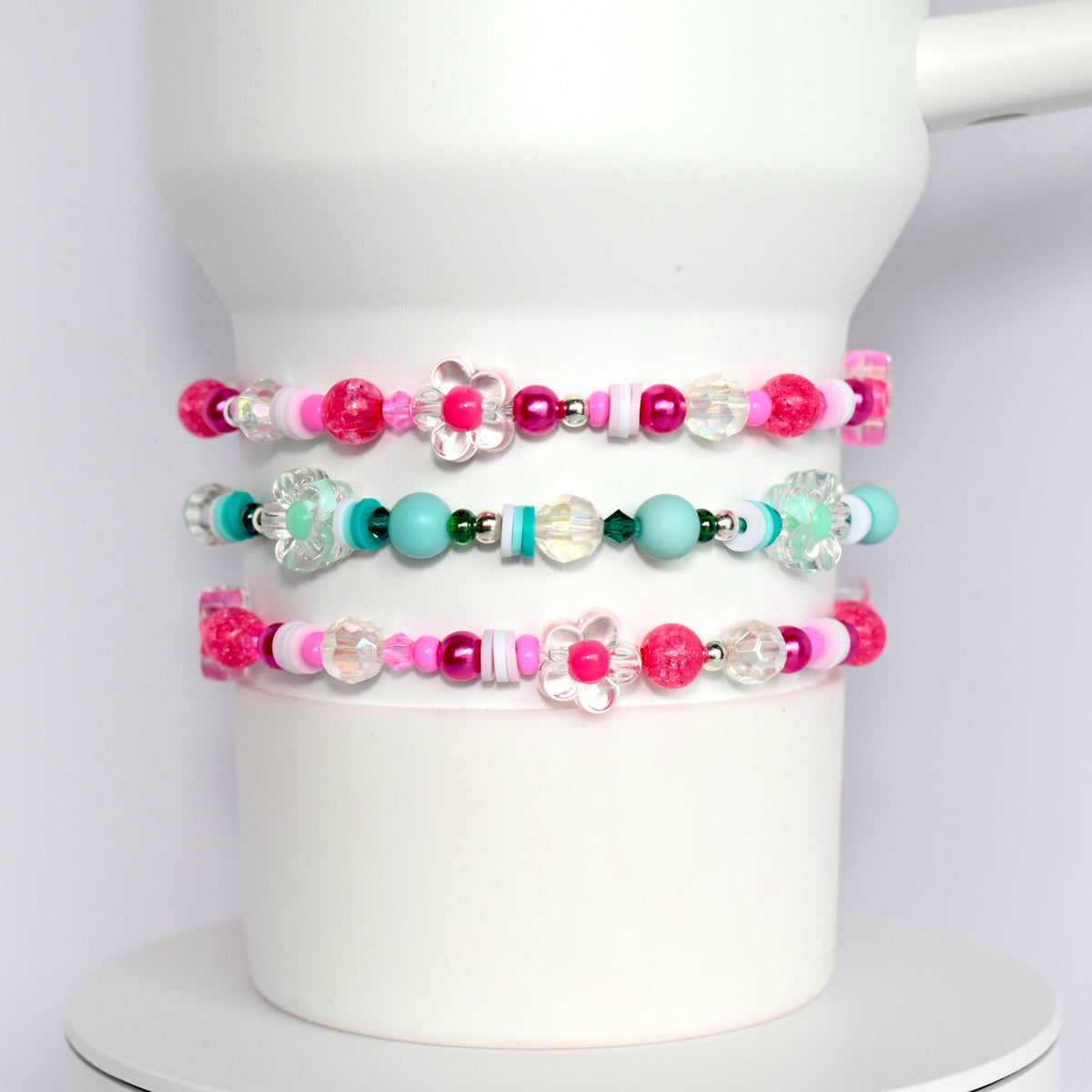 Three pink, white, and turquoise beaded bracelets with flower charms. The bracelets are stacked on top of each other.