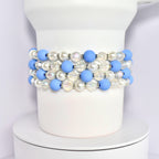 A set of four beaded bracelets around the base of a white Stanley tumbler. Each bracelet is made of blue matte beads, white pearl beads, and clear iridescent beads with silver spacer beads.