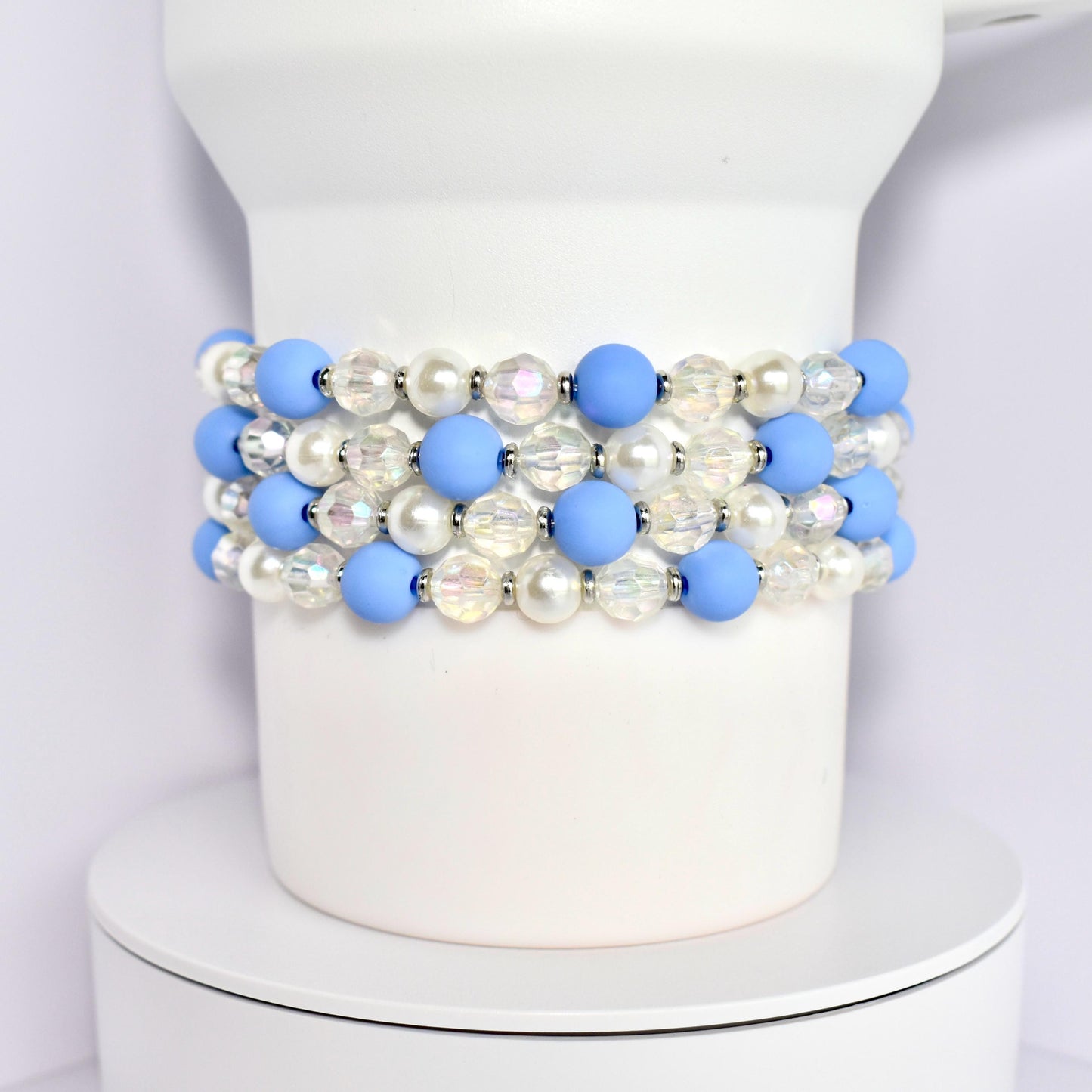 A set of four beaded bracelets around the base of a white Stanley tumbler. Each bracelet is made of blue matte beads, white pearl beads, and clear iridescent beads with silver spacer beads.