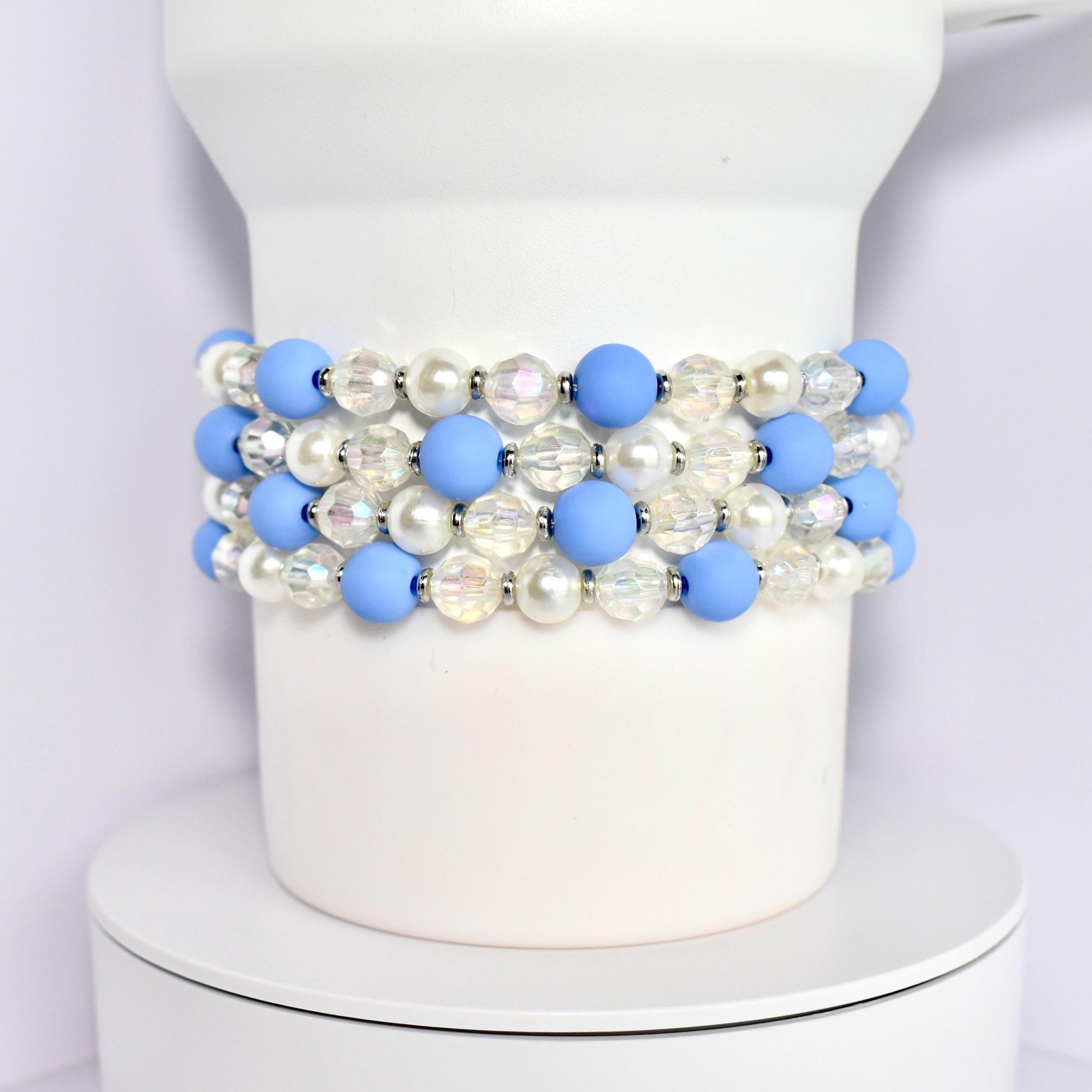 A set of four beaded bracelets around the base of a white Stanley tumbler. Each bracelet is made of blue matte beads, white pearl beads, and clear iridescent beads with silver spacer beads.