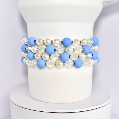 A set of four beaded bracelets around the base of a white Stanley tumbler. Each bracelet is made of blue matte beads, white pearl beads, and clear iridescent beads with silver spacer beads.