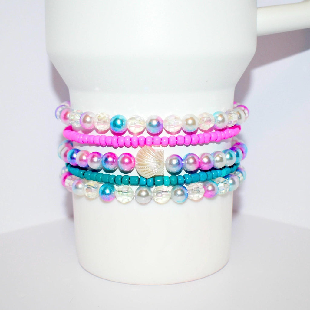 A stack of colorful tumbler bracelets wrapped around a white tumbler, featuring bright pink and teal seed beads alongside iridescent and pearl-like round beads. The center bracelet includes a pearlescent seashell charm, adding a beachy, playful touch to the vibrant and summery design.
