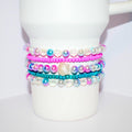 A stack of colorful tumbler bracelets wrapped around a white tumbler, featuring bright pink and teal seed beads alongside iridescent and pearl-like round beads. The center bracelet includes a pearlescent seashell charm, adding a beachy, playful touch to the vibrant and summery design.