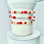 Three beaded bracelets with red, white, gold, and clear beads. One bracelet spells out the name "JACKI" in gold letters on white beads. The bracelets are displayed on a white cylindrical object.