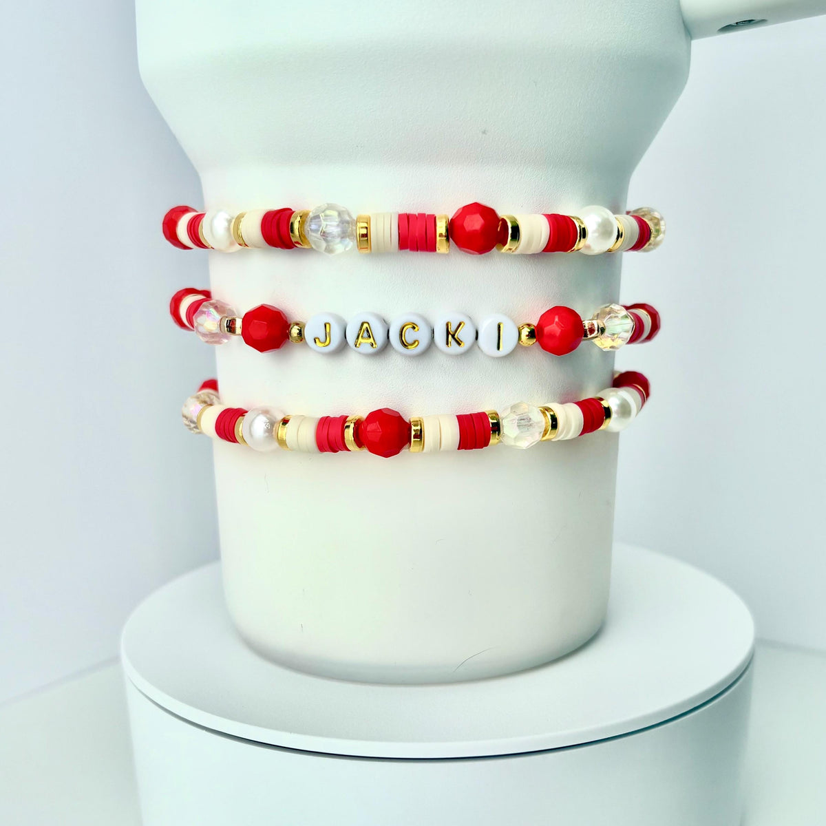 Three beaded bracelets with red, white, gold, and clear beads. One bracelet spells out the name "JACKI" in gold letters on white beads. The bracelets are displayed on a white cylindrical object.