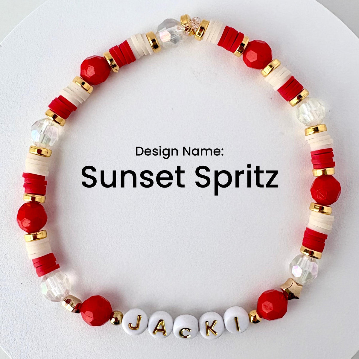 A beaded bracelet with the text "Sunset Spritz." The bracelet features red, white, gold, and clear beads. The name "JACKI" is spelled out in white letter beads. The bracelet is designed with a mix of round and disc-shaped beads.